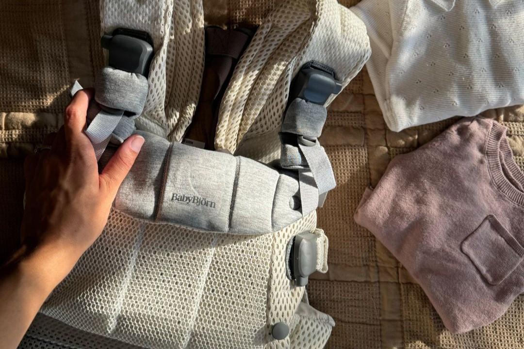 A hand holds a BabyBjörn baby carrier on a quilted surface beside a pink sweater and a white garment in soft lighting.