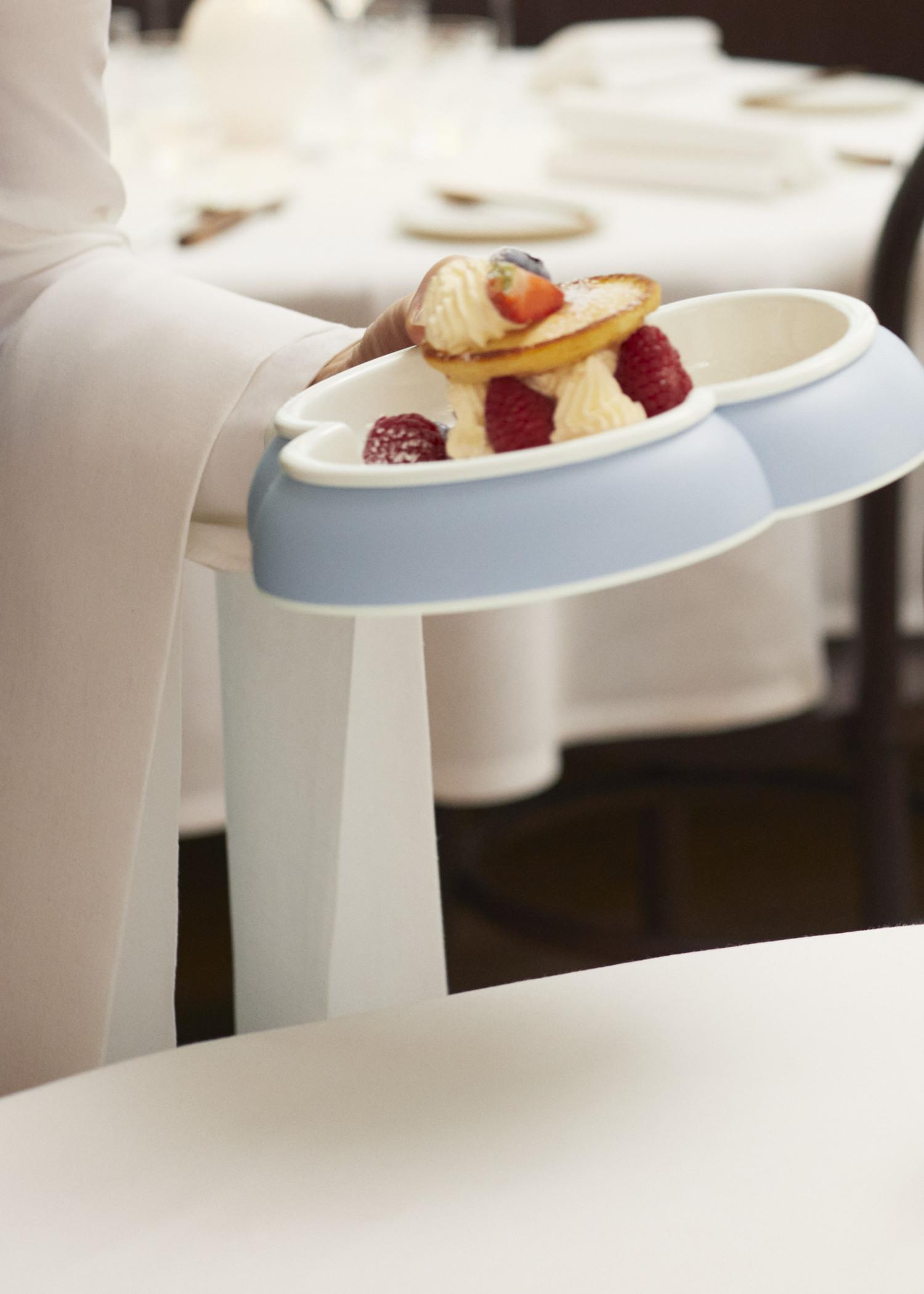 A person holds a blue tray with desserts topped with raspberries and strawberries, with a blurred dining setting in the background.