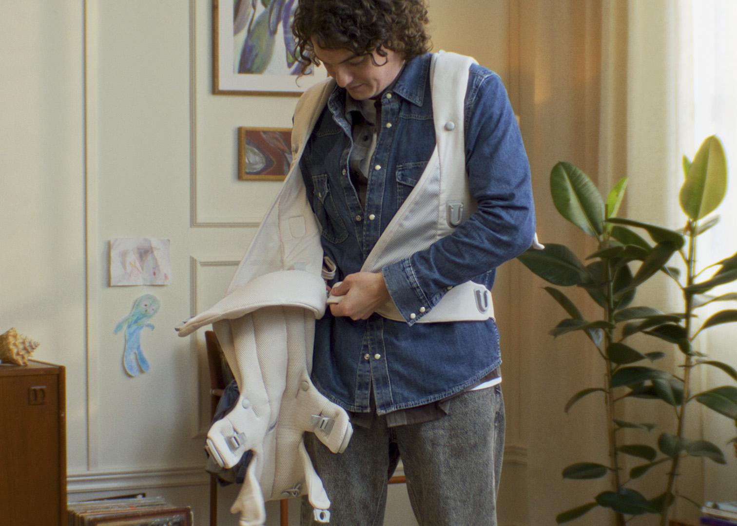 A person wearing a denim shirt is adjusting a baby carrier harness in a cozy room with a plant and artwork on the walls.