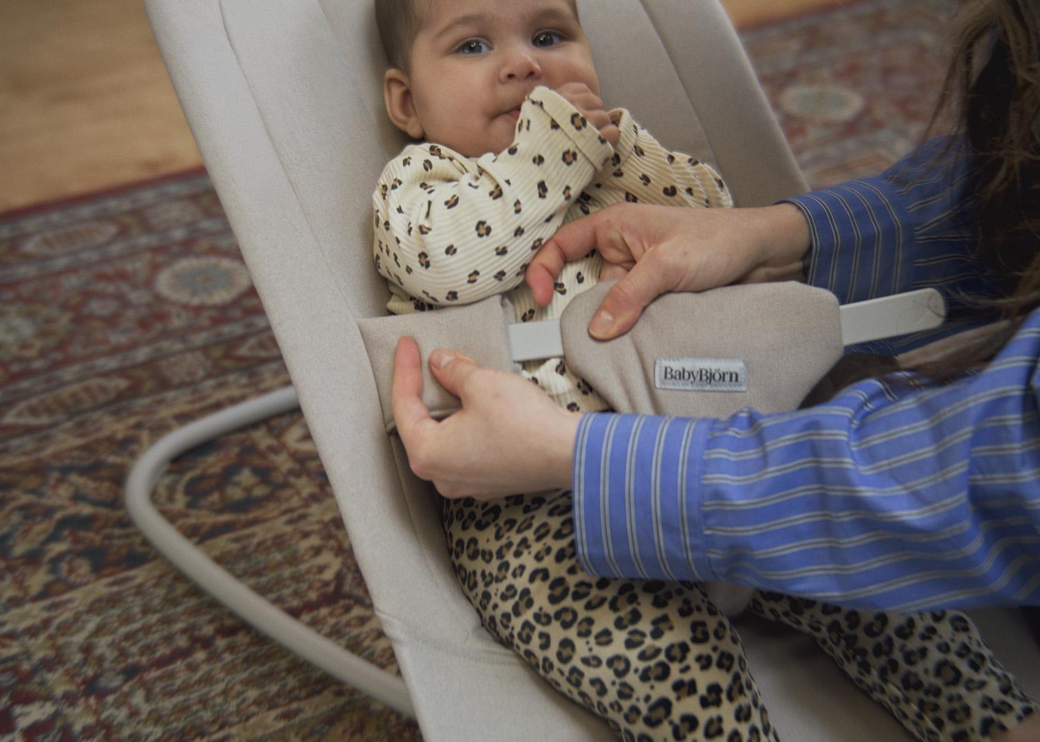 Baby in a bouncer with polka dot top and leopard print pants as an adult secures the safety harness.