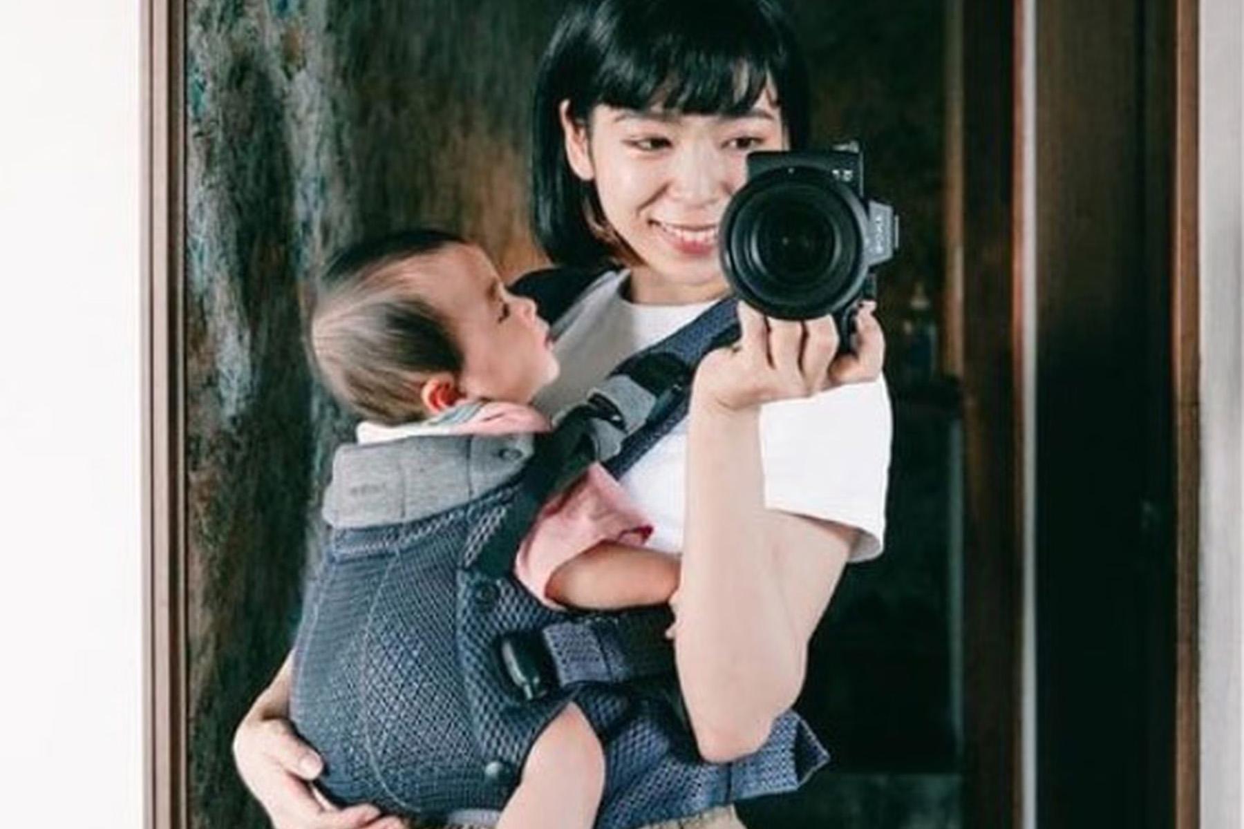 A woman smiles as she holds a camera, capturing a mirror selfie with a baby in a carrier on her chest.