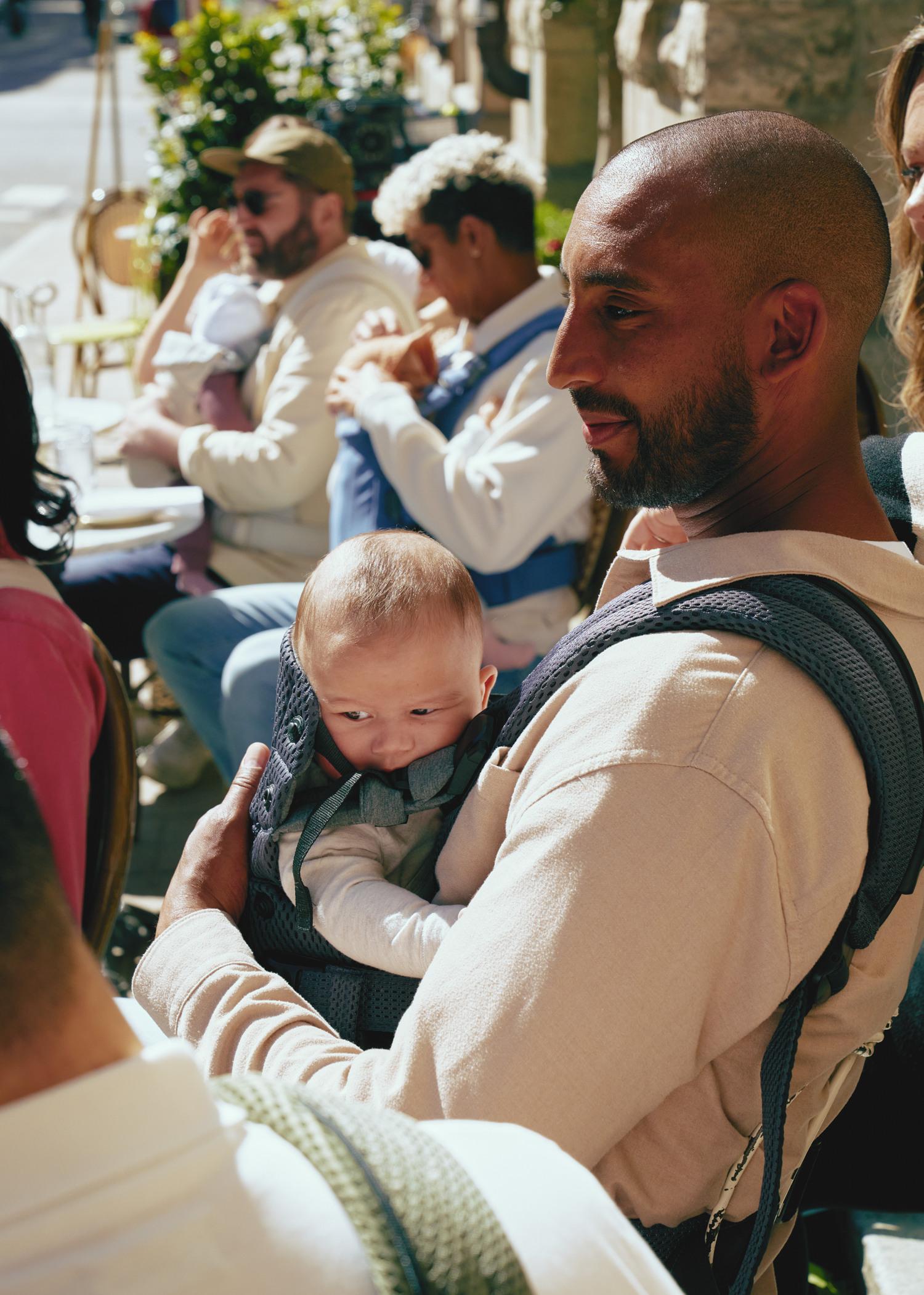 A man holds a baby in a carrier, surrounded by people sitting at an outdoor cafe. Sunlight casts shadows on the scene.