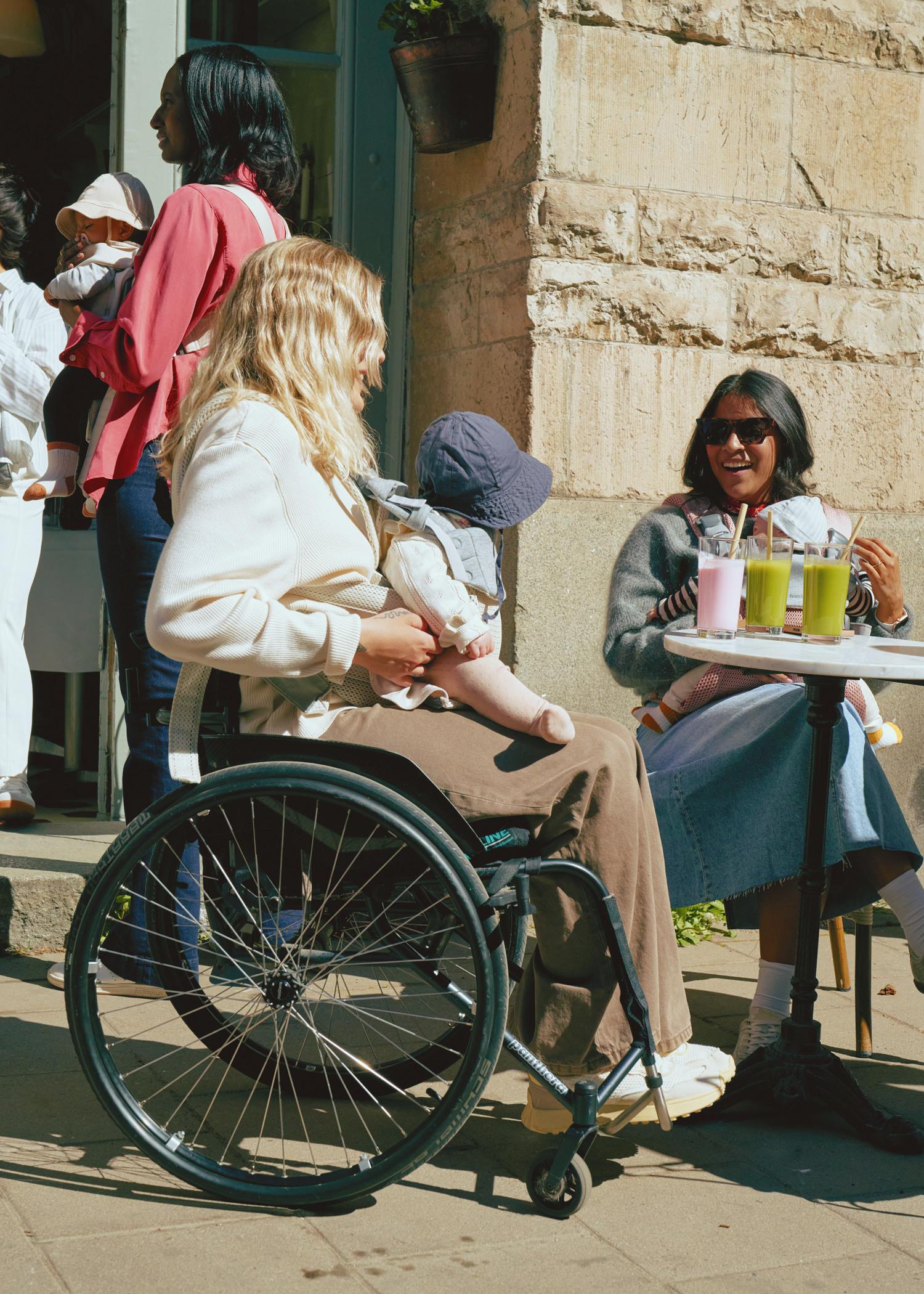 Woman in a wheelchair holding a baby, talking to another woman at a cafe table with colorful drinks; people and a baby in the background.