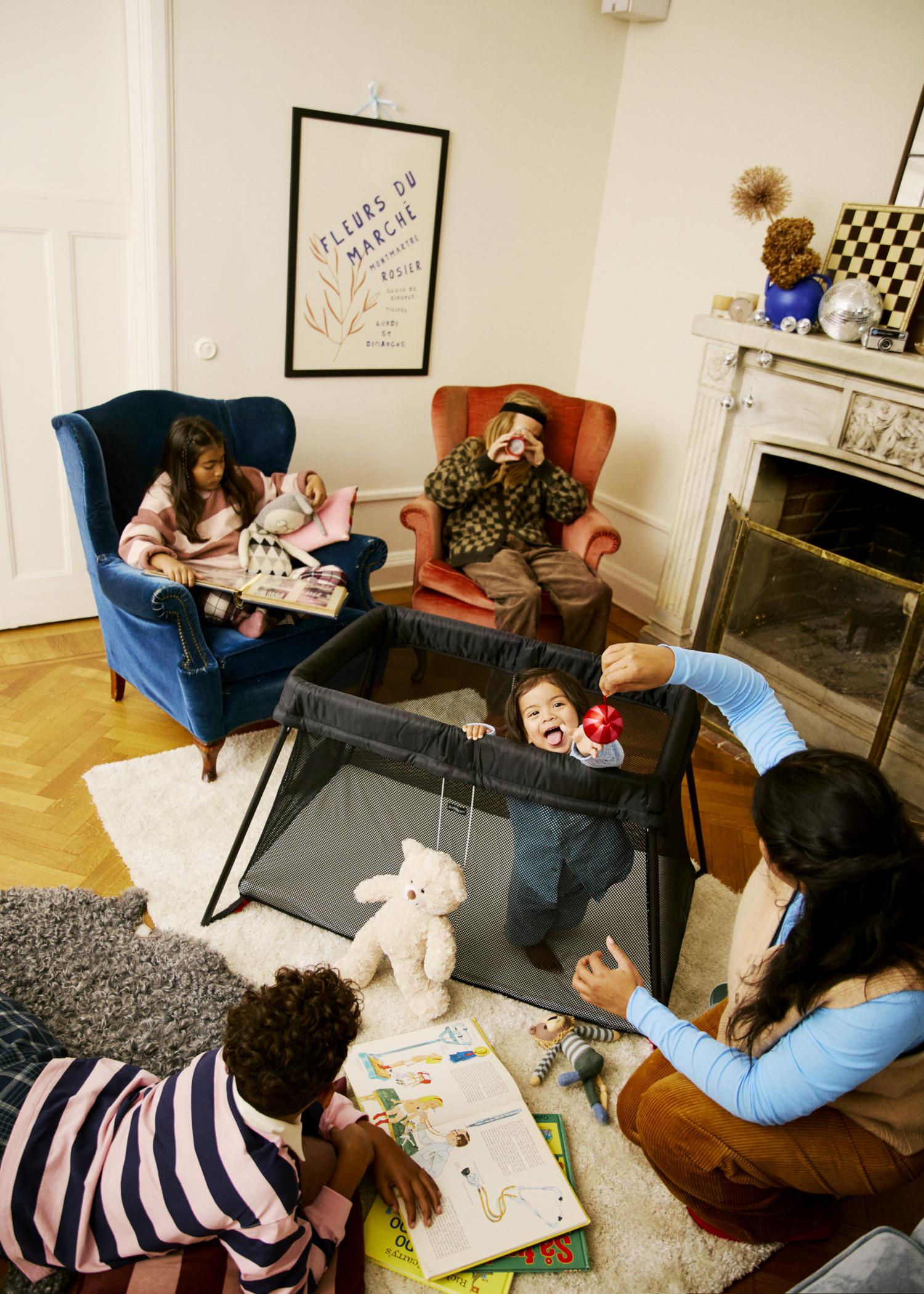 Children and adults in a cozy living room, playing and reading around a playpen with a baby, surrounded by books and toys.