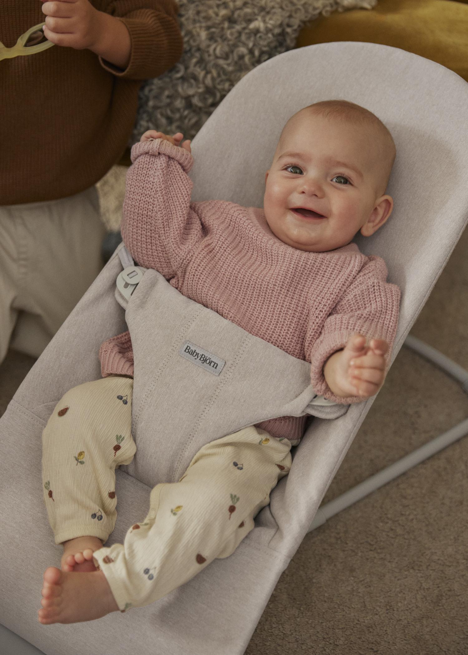 Smiling baby in a pink sweater and patterned pants sitting in a gray baby bouncer.