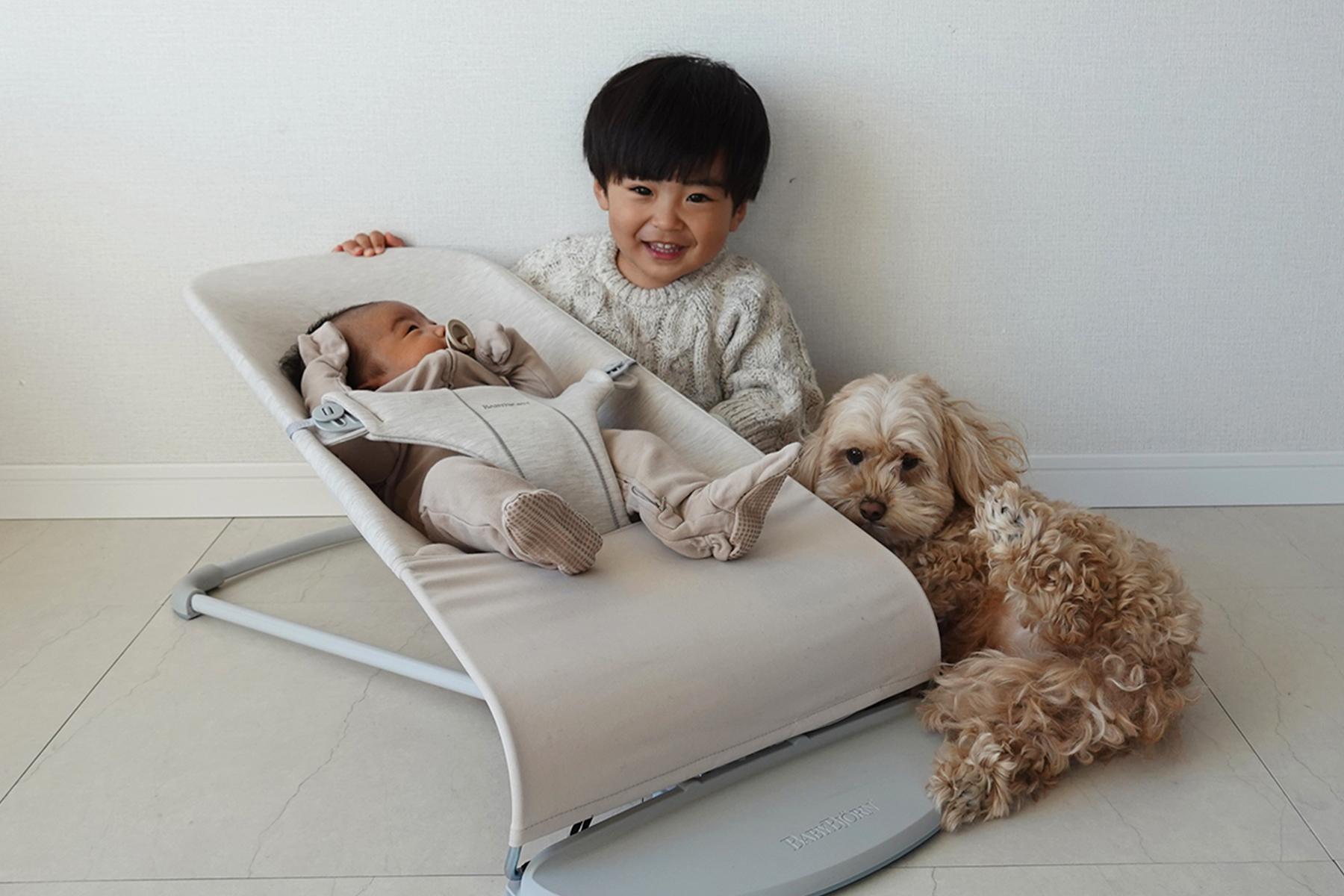 A young child smiles while sitting next to a baby in a bouncer. A fluffy dog lies beside them on a light-colored floor.