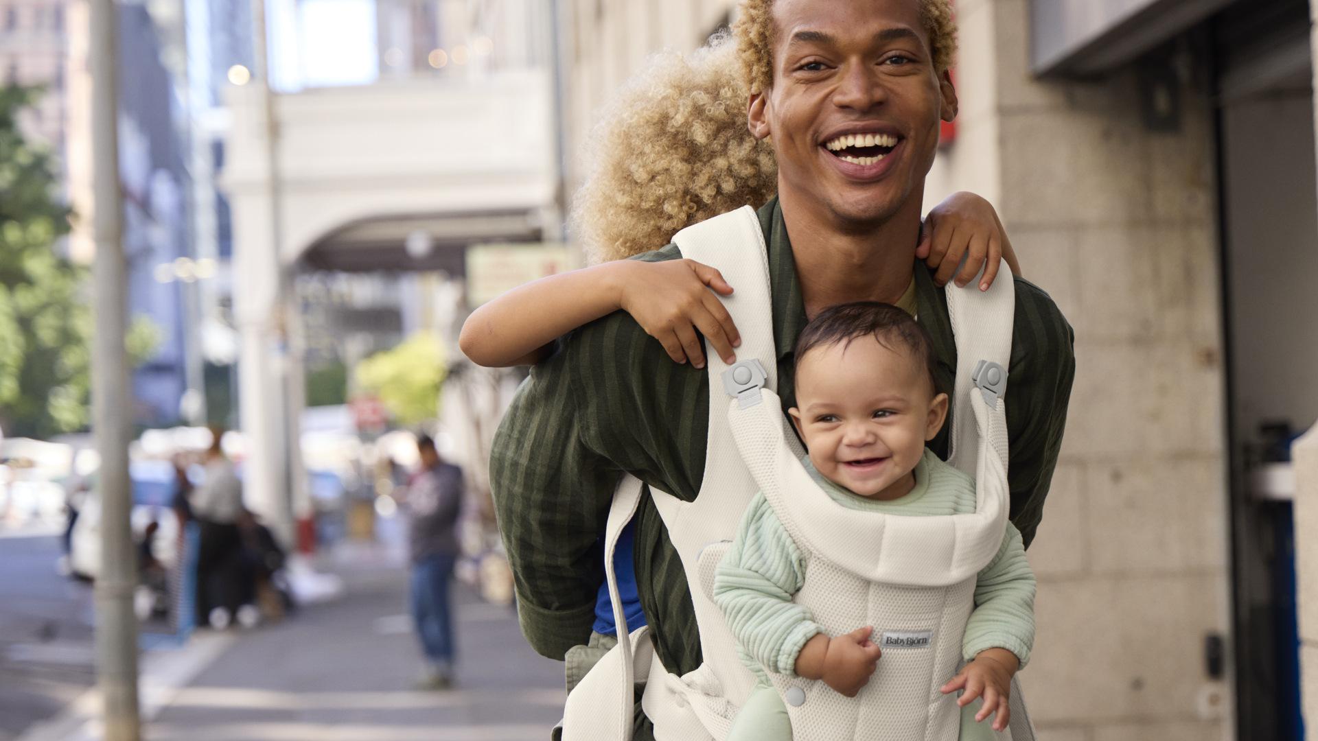 A smiling person in a city street carries a baby in a front carrier and a child on their back, both looking happy.