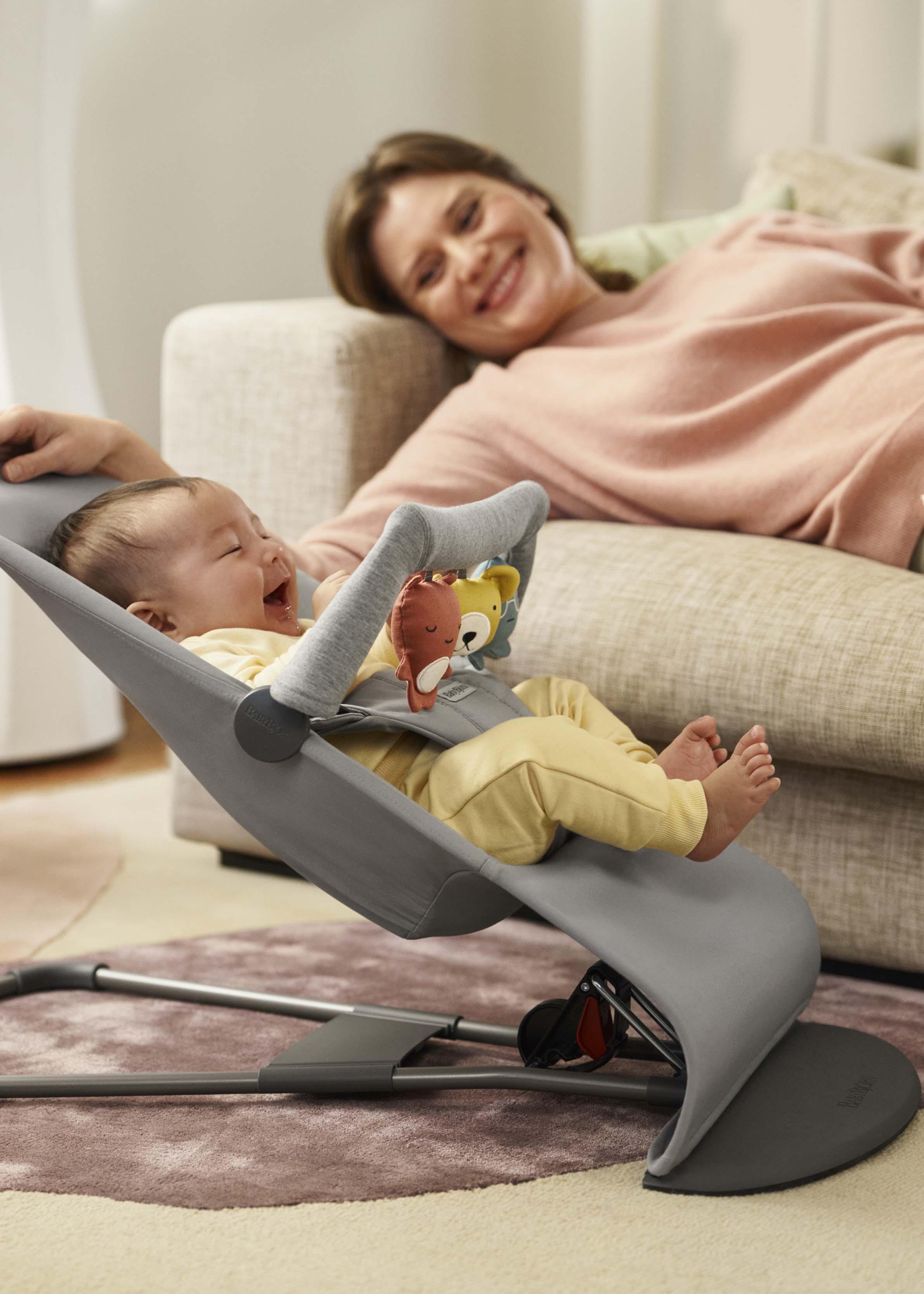 A smiling baby in a bouncer plays with toys, while a woman relaxes on a couch nearby.