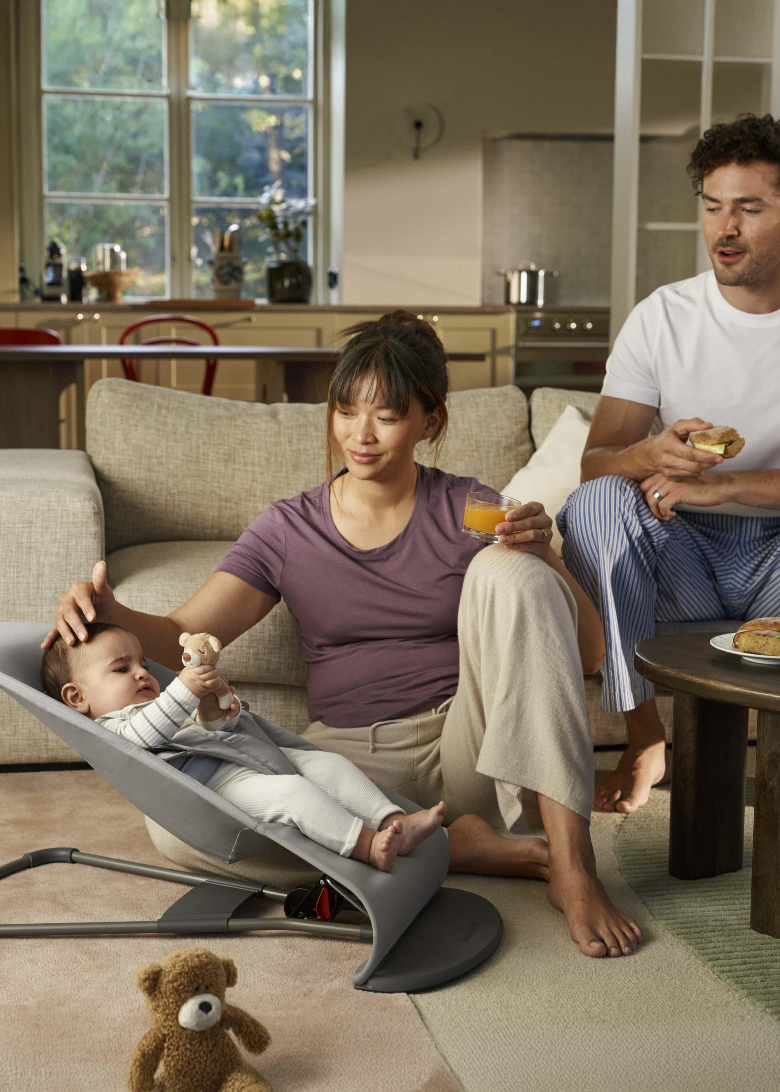 A couple sits on the floor of a cozy living room. The woman holds a smiling baby in a bouncer, while the man eats a pastry.